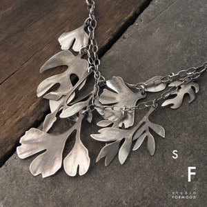 Op de afbeelding: Een zilveren ketting met meerdere bladeren als hangers. De bladeren zijn in een watervalvorm gerangschikt en met elkaar verbonden door een zilveren ketting. De ketting ligt op een donkergrijs oppervlak.