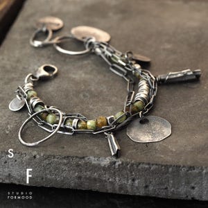Puede incluir: Una pulsera de plata con eslabones de cadena, cuentas verdes y marrones y peque&ntilde;os dijes circulares planos. La pulsera est&aacute; sobre una superficie gris oscuro. El texto "S F STUDIO FORMOOD" es visible.