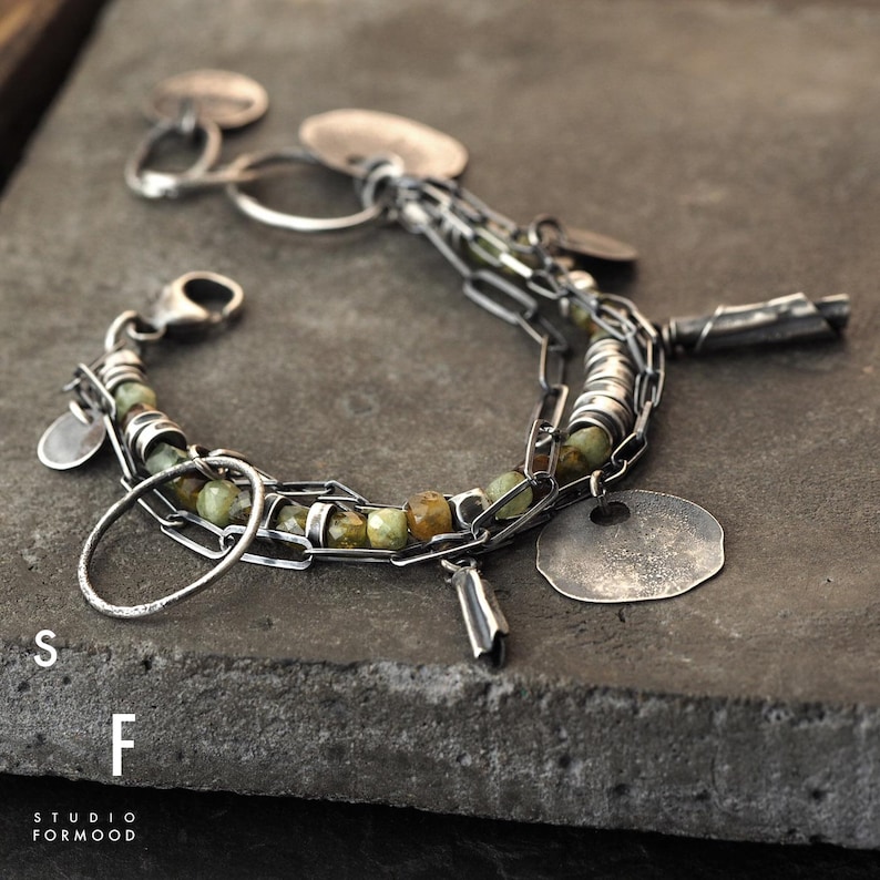Puede incluir: Una pulsera de plata con eslabones de cadena, cuentas verdes y marrones y peque&ntilde;os dijes circulares planos. La pulsera est&aacute; sobre una superficie gris oscuro. El texto "S F STUDIO FORMOOD" es visible.