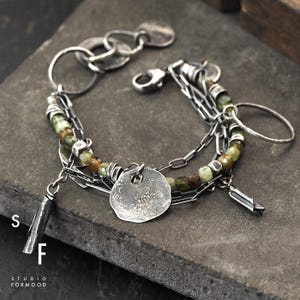Puede incluir: Una pulsera de plata con m&uacute;ltiples hilos, cadenas y dijes. La pulsera presenta una mezcla de texturas, incluyendo cuentas redondas en tonos verdes y marrones, y detalles de metal martillado. El cierre es un simple cierre de gancho. El texto "S F STUDIO FORMOOD" es visible.