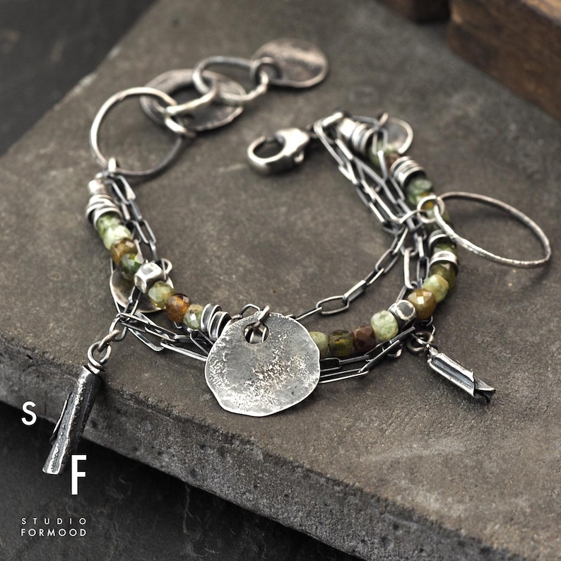 Puede incluir: Una pulsera de plata con m&uacute;ltiples hilos, cadenas y dijes. La pulsera presenta una mezcla de texturas, incluyendo cuentas redondas en tonos verdes y marrones, y detalles de metal martillado. El cierre es un simple cierre de gancho. El texto "S F STUDIO FORMOOD" es visible.