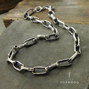 Puede incluir: Un collar plateado con un dise&ntilde;o de cadena texturizada &uacute;nico. Los eslabones tienen forma irregular, lo que le da al collar una apariencia artesanal. El collar se muestra sobre una superficie gris, con el logotipo del estudio, "FORMood", visible en la esquina inferior derecha.