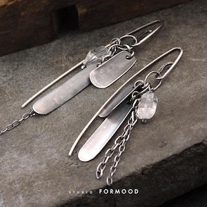 Peut inclure: Une paire de boucles d'oreilles en argent au design unique. Chaque boucle d'oreille comporte une longue barre fine en argent, une petite étiquette rectangulaire et une perle de cristal transparente. Une chaîne délicate pend d'une boucle d'oreille.