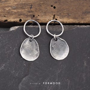 Pode incluir: Um par de brincos de tom prateado com um aro circular preso a um pendente irregular em forma de disco texturizado. O metal tem um acabamento escuro e oxidado com uma superfície rugosa. O texto STUDIO FORMOOD é visível na parte inferior.