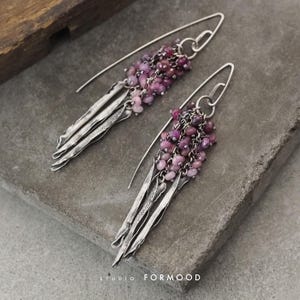 Può includere: Un paio di orecchini in argento con un grappolo di piccole pietre preziose rosa e elementi argentati allungati e strutturati. Gli orecchini hanno un design pendente e sono esposti su una superficie grigia. La parola "FORMOOD" è stampata in basso a destra.