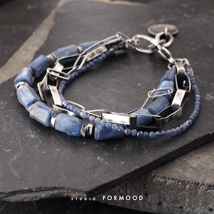 Könnte beinhalten: Ein mehrreihiges Armband mit zylindrischen blauen Kyanitperlen, einer silberfarbenen Metallkette mit rechteckigen Gliedern und einem feinen Strang aus kleinen facettierten blauen Perlen. Das Schmuckstück hat einen silberfarbenen Verschluss und einen kleinen runden Anhänger.