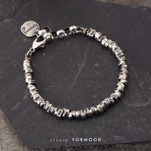 Puede incluir: Una pulsera de plata compuesta por numerosas cuentas metálicas cilíndricas y texturizadas. La pulsera cuenta con un cierre de mosquetón, una cadena de extensión corta y una pequeña etiqueta metálica redonda grabada con el texto formood 925.