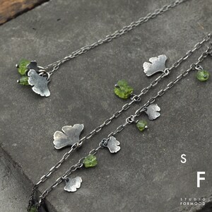 Peut inclure: Deux cha&icirc;nes en argent avec des accents de pierres pr&eacute;cieuses vertes et des breloques en forme de feuilles de ginkgo en argent. Les cha&icirc;nes sont superpos&eacute;es et les breloques pendent. Le texte "Studio Formwood" et les lettres "S" et "F" sont visibles dans le coin inf&eacute;rieur droit de l'image.