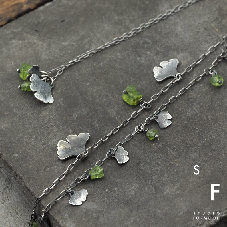 Peut inclure: Deux cha&icirc;nes en argent avec des accents de pierres pr&eacute;cieuses vertes et des breloques en forme de feuilles de ginkgo en argent. Les cha&icirc;nes sont superpos&eacute;es et les breloques pendent. Le texte "Studio Formwood" et les lettres "S" et "F" sont visibles dans le coin inf&eacute;rieur droit de l'image.