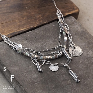 Könnte beinhalten: Eine silberne Halskette mit mehreren Strängen und baumelnden Anhängern. Die Halskette verfügt über verschiedene Kettenstile, kleine runde Perlen und flache kreisförmige Anhänger. Das Design umfasst rechteckige und zylindrische Silberelemente. Der Text "S F STUDIO FORMOOD" ist sichtbar.