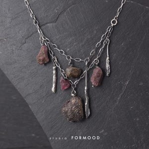Pode incluir: Um colar de corrente de prata com vários elementos pendentes, incluindo pedras brutas e irregulares cor-de-rosa e castanhas e barras de metal prateado texturizadas. O texto STUDIO FORMOOD é visível na parte inferior da imagem.