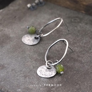 Puede incluir: Pendientes de plata con un disco martillado y una cuenta de piedra preciosa verde. Los pendientes tienen un diseño de aro ovalado y un cierre de poste. El texto "studio FORMOOD" está en la parte inferior.