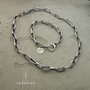 Puede incluir: Un conjunto de collar y pulsera de plata con un dise&ntilde;o de eslabones de cadena. El collar y la pulsera se muestran sobre una superficie gris. La pulsera tiene un peque&ntilde;o dije redondo con la palabra "formood".