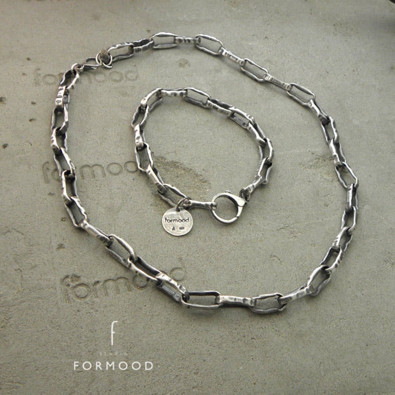 Puede incluir: Un conjunto de collar y pulsera de plata con un dise&ntilde;o de eslabones de cadena. El collar y la pulsera se muestran sobre una superficie gris. La pulsera tiene un peque&ntilde;o dije redondo con la palabra "formood".