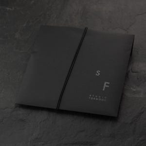 Puede incluir: Un sobre negro con cierre de banda el&aacute;stica negra. Las letras "S" y "F" est&aacute;n impresas en la parte delantera, junto con las palabras "STUDIO FORMOOD". El sobre est&aacute; hecho de un material mate.