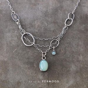 Puede incluir: Un collar de plata con un colgante ovalado de piedra preciosa azul claro y una piedra preciosa redonda más pequeña. El collar presenta múltiples capas de cadena y eslabones ovalados texturizados. El texto "STUDIO FORMOOD" está en la parte inferior.