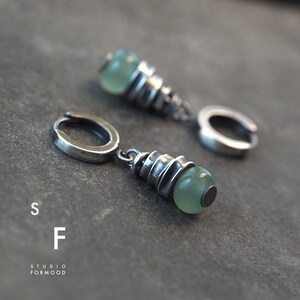 May include: A pair of silver hoop earrings with a green stone pendant. The pendant is made of stacked silver rings with a green stone at the bottom.