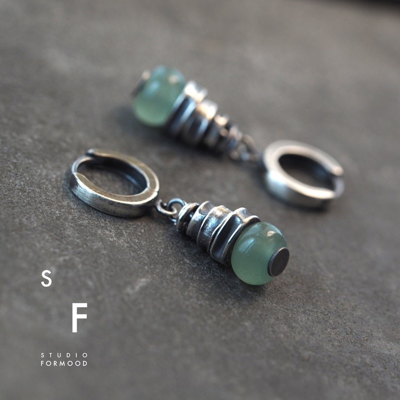 May include: A pair of silver hoop earrings with a green stone pendant. The pendant is made of stacked silver rings with a green stone at the bottom.