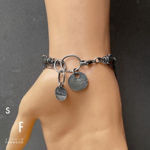 Puede incluir: Una pulsera plateada con un dise&ntilde;o de eslabones y dos colgantes circulares. Un colgante es de forma ovalada peque&ntilde;a con la palabra "formood" grabada. El otro colgante es de forma redonda m&aacute;s grande con superficie texturizada.