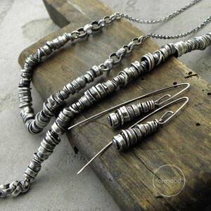 May include: A set of silver jewellery featuring a necklace, bracelet, and earrings. The jewellery is made of small, stacked silver rings. The jewellery is displayed on a wooden surface.
