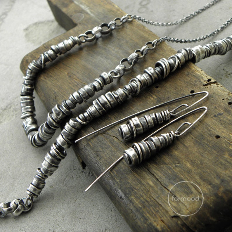 May include: A set of silver jewellery featuring a necklace, bracelet, and earrings. The jewellery is made of small, stacked silver rings. The jewellery is displayed on a wooden surface.