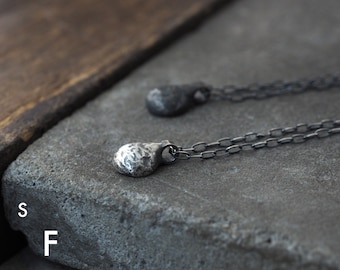 May include: Two silver pendant necklaces with textured, teardrop-shaped pendants. The pendants hang from delicate chain necklaces. The image is set on a gray stone surface. The text "SF STUDIO FORMOOD" is visible in the lower left corner.