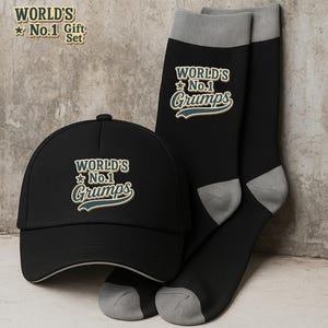 Pode incluir: Um conjunto de presente com um boné de beisebol preto e um par de meias pretas com detalhes cinzentos. O boné e as meias apresentam o texto "WORLD'S No.1 Grumps" em estilo vintage.