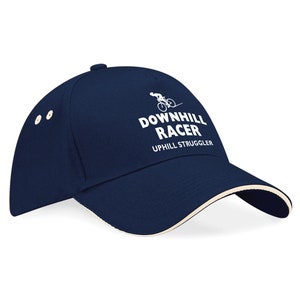 Könnte beinhalten: Eine marineblaue Baseballkappe mit einem weißen gestickten Design, auf dem "Downhill Racer" und "Uphill Struggler" steht. Das Design enthält auch eine Silhouette einer Person, die Fahrrad fährt.