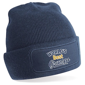 Könnte beinhalten: Eine marineblaue Beanie-Mütze mit umgeschlagenem Saum. Die Mütze hat einen rechteckigen Aufnäher mit den Worten "WORLD'S Best Grumps" in weißer und goldener Schrift. Die Mütze ist aus Strickmaterial.