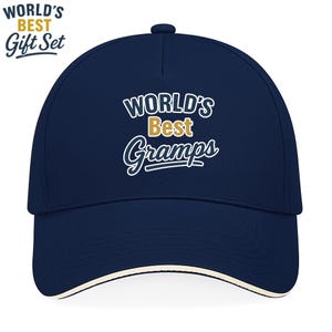 Könnte beinhalten: Eine marineblaue Baseballkappe mit dem Text "WORLD'S BEST Gramps" in weißer und goldener Schrift. Die Kappe hat einen gebogenen Schirm und einen weißen Besatz. Die Worte "WORLD'S BEST Gift Set" sind ebenfalls sichtbar.