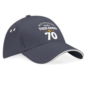 May include: A navy blue baseball cap with a white brim and a white embroidered design that says "Not everyone looks this good at 70".