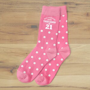 K&ouml;nnte beinhalten: Ein Paar rosa Socken mit wei&szlig;en Punkten. Die Socken haben ein wei&szlig;es Textdesign mit der Aufschrift "Not everyone looks this good at 21".