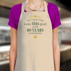 Pode incluir: Avental de cozinha bege com um design verde e dourado. O texto diz "Nem todos os cozinheiros ficam t&atilde;o bem depois de 80 anos na cozinha".