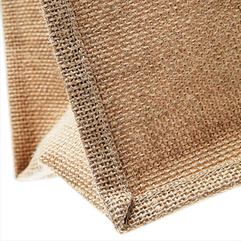 Puede incluir: Primer plano de la costura de una bolsa de mano de yute marr&oacute;n natural. La tela est&aacute; tejida con una textura rugosa.