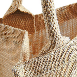 Puede incluir: Primer plano de una bolsa de mano de arpillera marr&oacute;n con un asa gruesa tejida. La bolsa est&aacute; hecha de fibras naturales y tiene un aspecto r&uacute;stico y terroso.