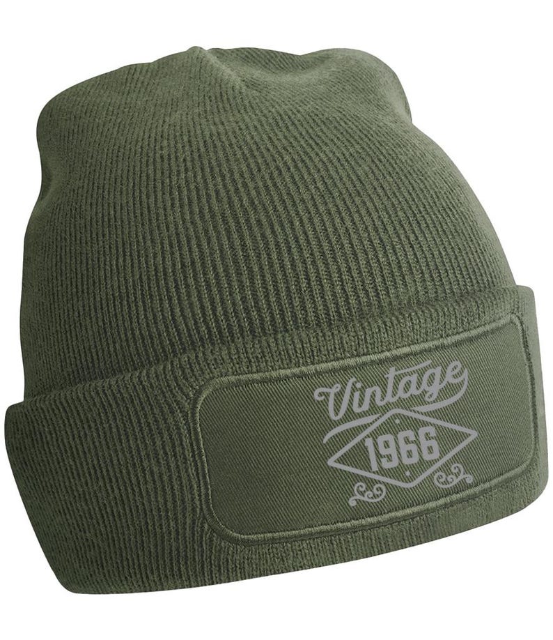 May include: An olive green knitted beanie hat with a rectangular patch. The patch displays the word "Vintage" above the year "1966" in a diamond shape, in light grey. A winter hat.