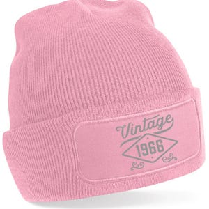 May include: A pale pink beanie hat with a rectangular patch. The patch displays the word "Vintage" above the year "1966" in a diamond shape, in grey. The hat is made of a knitted material.