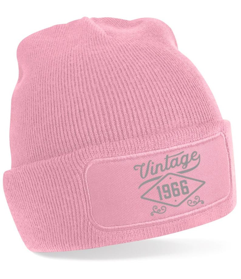 May include: A pale pink beanie hat with a rectangular patch. The patch displays the word "Vintage" above the year "1966" in a diamond shape, in grey. The hat is made of a knitted material.
