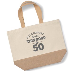May include: A beige canvas tote bag with brown jute trim. The bag has brown straps and features the text "NOT EVERYONE looks THIS GOOD at 50" in gray.