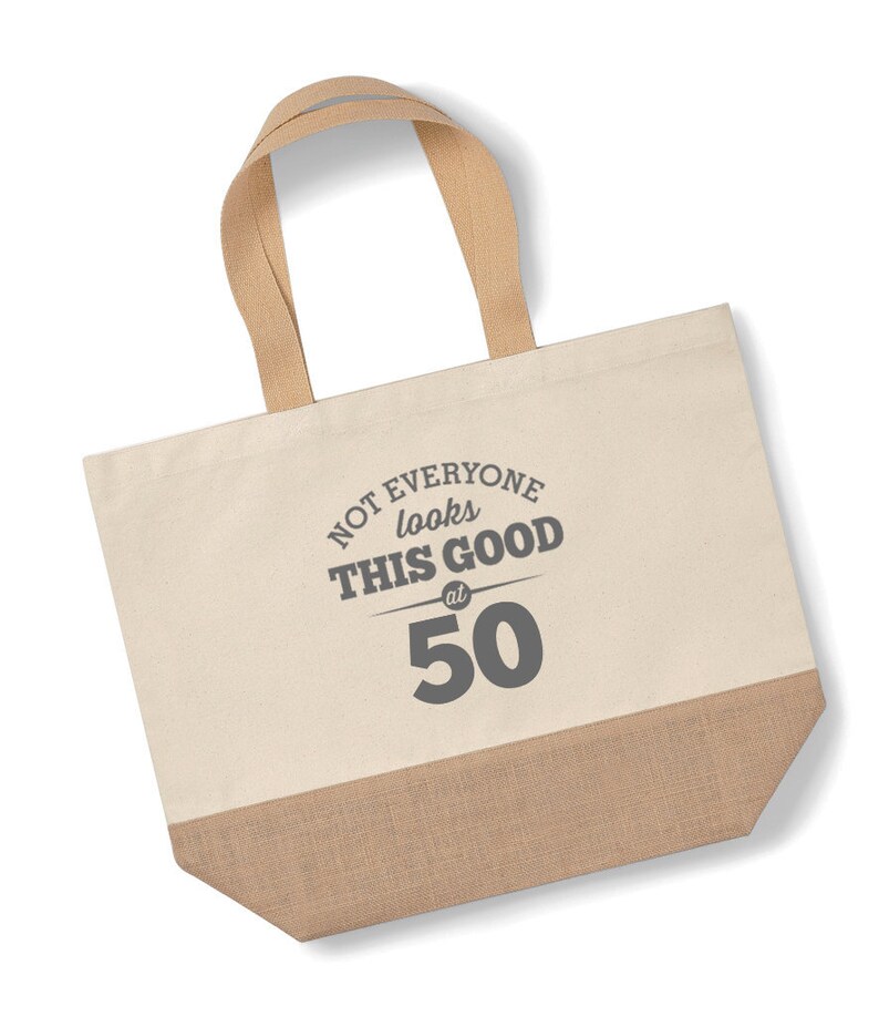 May include: A beige canvas tote bag with brown jute trim. The bag has brown straps and features the text "NOT EVERYONE looks THIS GOOD at 50" in gray.