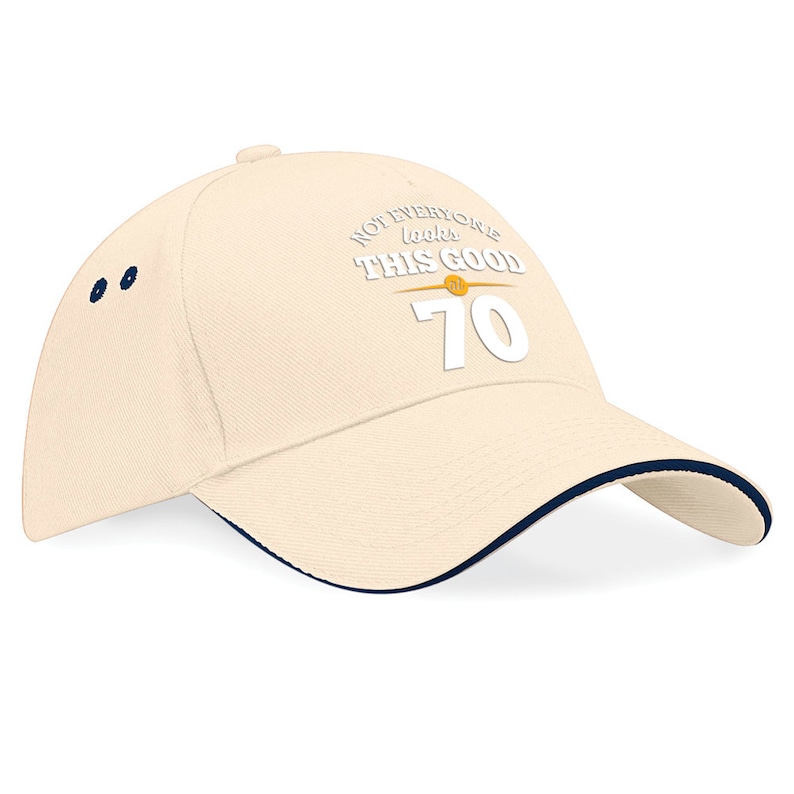 May include: A beige baseball cap with a navy blue brim. The front of the cap has white text that reads "Not everyone looks this good at 70".