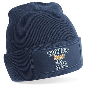 May include: A navy blue beanie hat with a folded brim. The hat has a rectangular patch with the words "WORLD'S Best Pa" in white and gold lettering. The hat is made of a soft, knitted material.