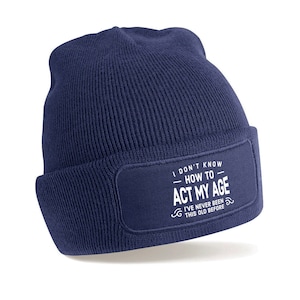 May include: A navy blue beanie with a white patch that reads "I don't know - how to - act my age. I've never been this old before."