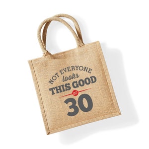 Puede incluir: Una bolsa de tela de yute marr&oacute;n con el texto "Not everyone looks this good at 30" impreso en ella. El texto est&aacute; en negro y rojo.