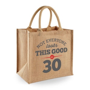 Puede incluir: Una bolsa de tela de yute marr&oacute;n con el texto "Not everyone looks this good at 30" impreso en gris y rojo.