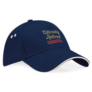 May include: A navy blue baseball cap with a white brim. The cap has gold embroidery that reads "Officially Retired" and a red banner that reads "Not My Problem".