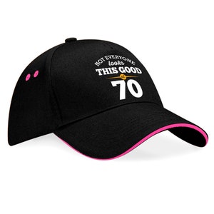 May include: Black baseball cap with a pink brim. The front of the cap has white text that reads "Not everyone looks this good at 70".