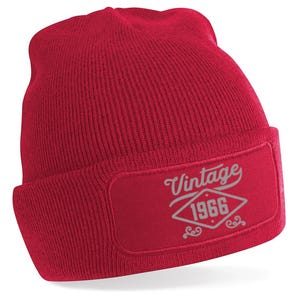 May include: A red knitted beanie hat with a turned-up brim. The hat has a rectangular patch with the text "Vintage 1966" in a decorative font. The beanie is made of a soft, ribbed fabric.