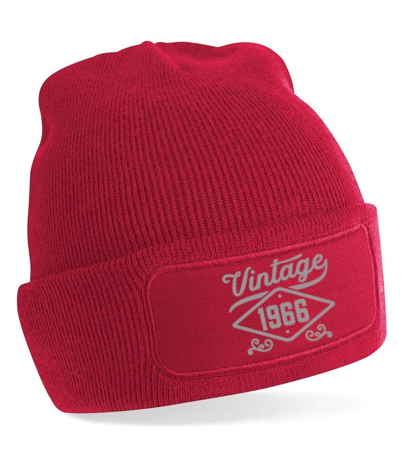 May include: A red knitted beanie hat with a turned-up brim. The hat has a rectangular patch with the text "Vintage 1966" in a decorative font. The beanie is made of a soft, ribbed fabric.