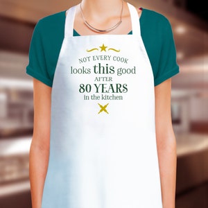 Pode incluir: Avental branco com o texto "Not every cook looks this good after 80 years in the kitchen" em letras verdes e douradas.